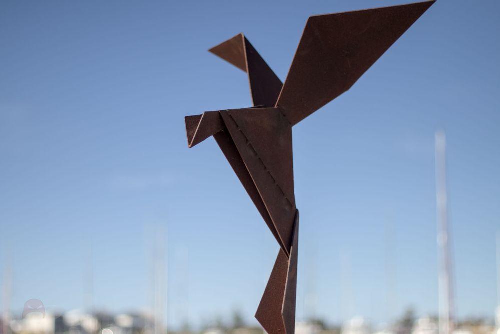 A rusted steel stylised bird with its wings swept back as if coming into land. It is done in an origami style