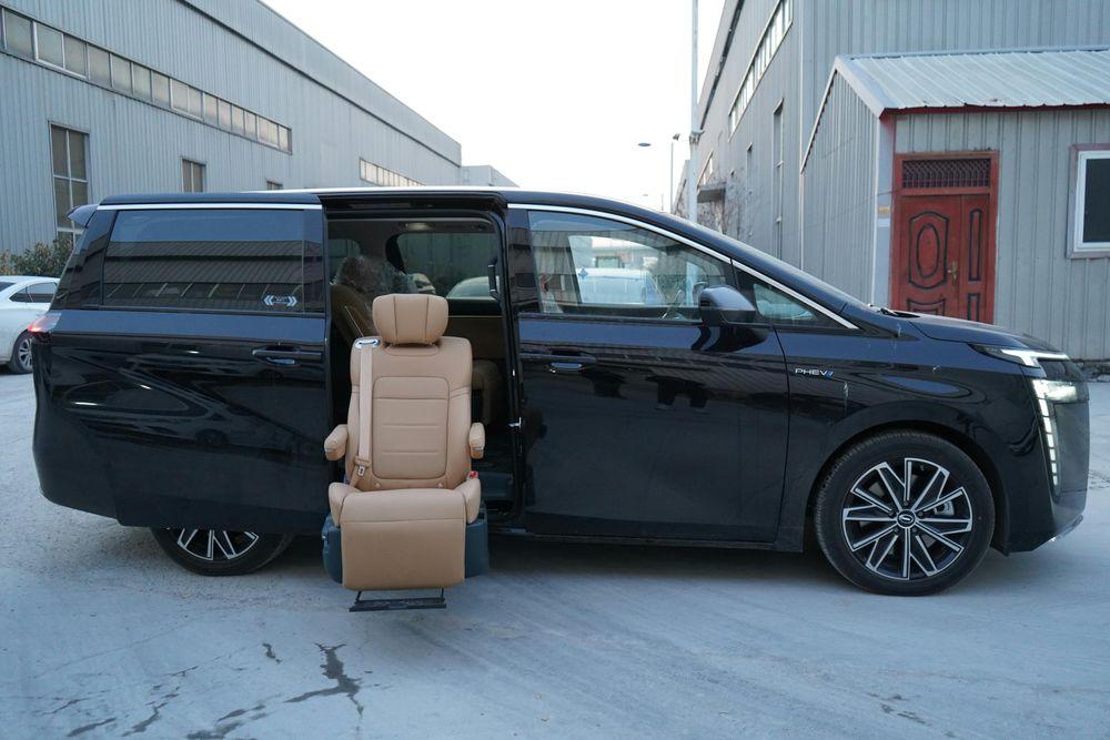 A black van sits in a grey industrial compound. Its side door is open and a beige chair has swivelled out