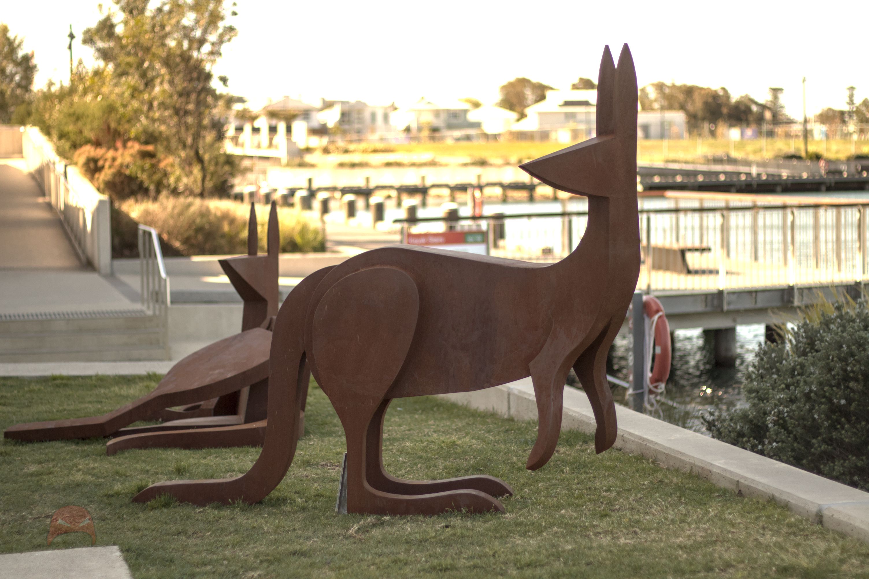 Two rust red kangaroo sculptures stand on grass. The one closest to the viewer is standing facing right but looking over its shoulder to the left. In the backgroun we can see the other sculpture laying on the ground, as if at rest